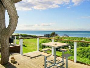 Une terrasse en bois avec une table et des bancs, donnant sur une pelouse et la mer.