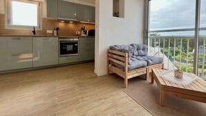 A modern kitchen with grey cabinets, a wooden countertop, and a built-in oven. There's a grey upholstered bench with a tray table and a wicker basket. A large window offers a view of greenery outside. - 47 Reflets- 4p-wifi-parking-vue mer (Quiberon)