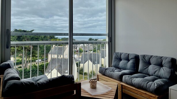 A balcony with a view of the city, a grey sofa, and a hanging light fixture. - 47 Reflets- 4p-wifi-parking-vue mer (Quiberon)