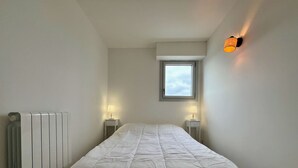 A small, neatly made room with a bed, two bedside tables with lamps, a radiator, and a window with a view of the sky. - 47 Reflets- 4p-wifi-parking-vue mer (Quiberon)