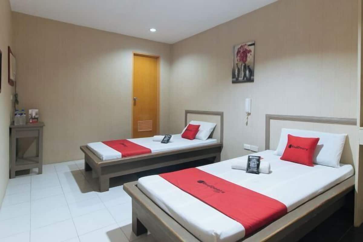 Ein Hotelzimmer mit zwei Betten, jeweils mit roter Decke und weißen Kissen. Eine Holztür, ein kleiner Tisch mit Lampe und ein Bild an der Wand sind ebenfalls vorhanden.