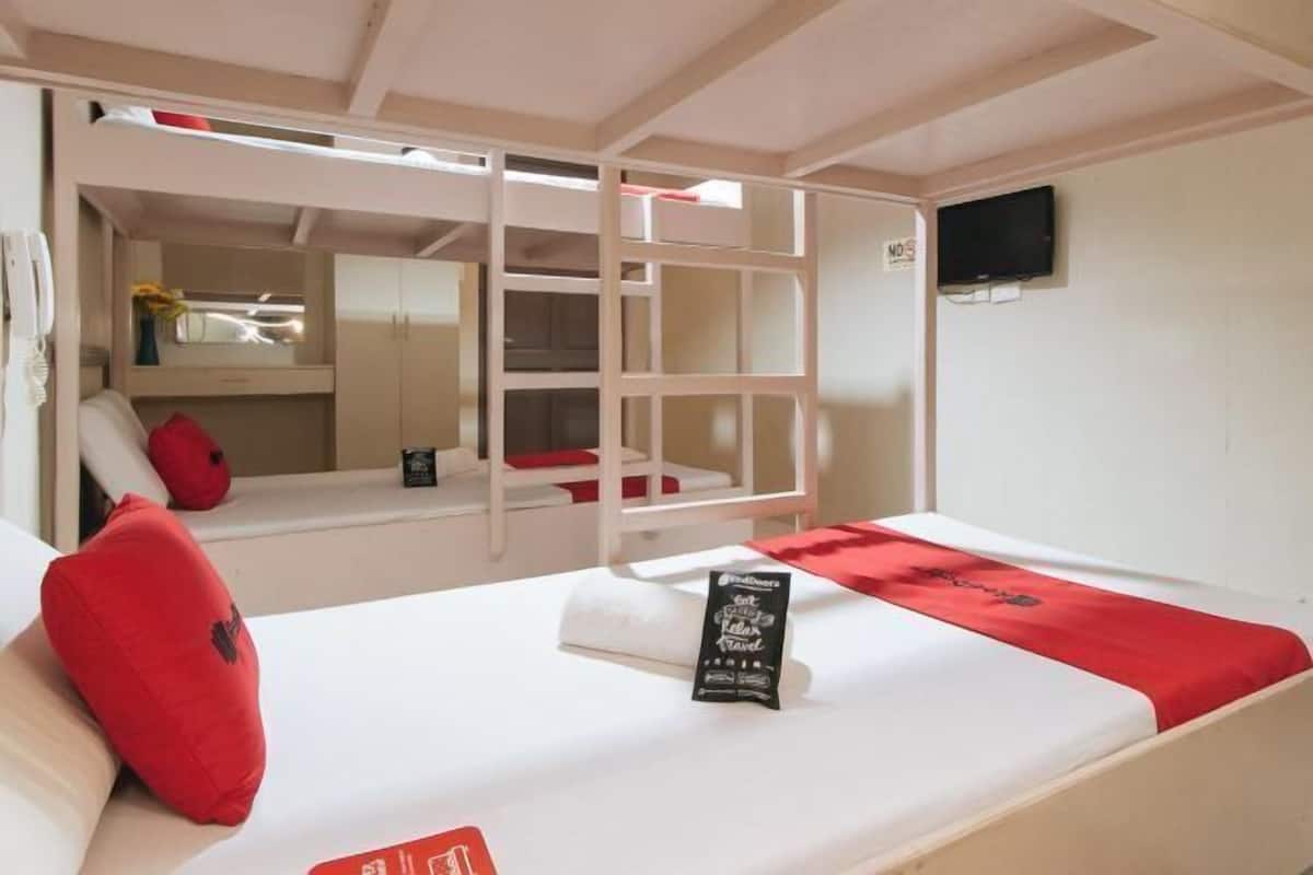 Ein Schlafraum mit Etagenbetten, einem Fernseher an der Wand, einem kleinen Tisch mit roter Tischdecke und einem roten Kissen auf dem Bett.
