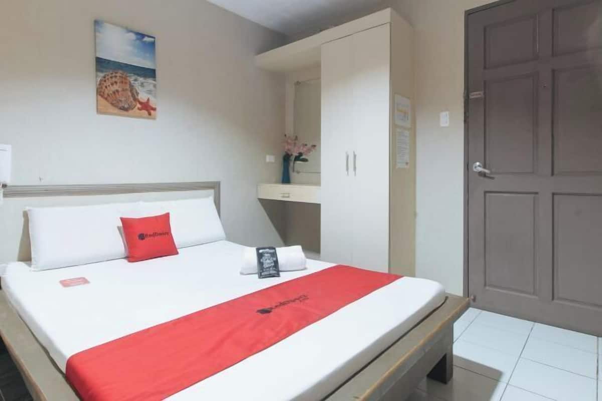 Ein Hotelzimmer mit einem Bett, einer roten Decke, einem Kopfkissen, einem Nachttisch, einem Spiegel und einer Tür.