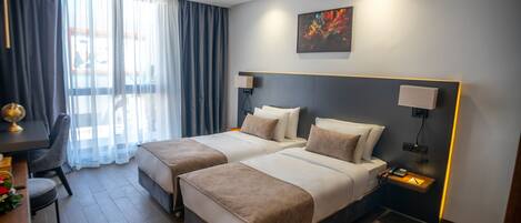 Ein Hotelzimmer mit zwei Betten, einem Schreibtisch und einem Sessel. Es gibt ein Fenster mit Vorhängen, ein Bild an der Wand und eine Lampe auf dem Nachttisch.