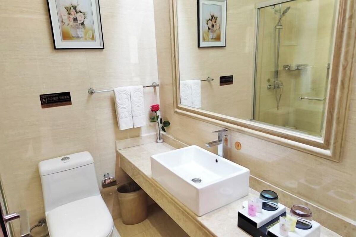 A modern bathroom with a white sink, toilet, and shower. There are framed pictures on the wall and a small trash bin.