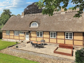 Une maison traditionnelle à toit de chaume, avec des fondations en pierre, un petit porche à l’avant et un patio aménagé avec du mobilier d’extérieur.