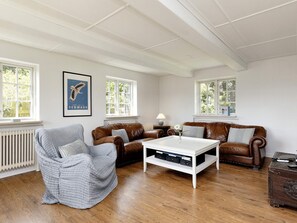 Un salon avec un canapé en cuir marron, un fauteuil gris et une table basse blanche. Il y a deux fenêtres à cadres blancs, un radiateur et un tableau encadré au mur.