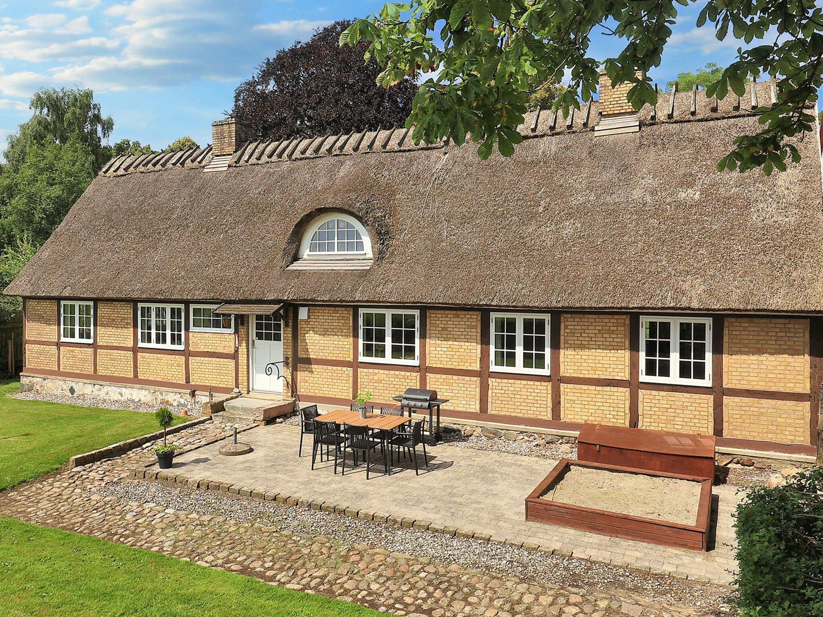 Ein traditionelles Haus mit einem Strohdach, einem Steinfundament, einer kleinen Veranda und einer Terrasse mit Gartenmöbeln.