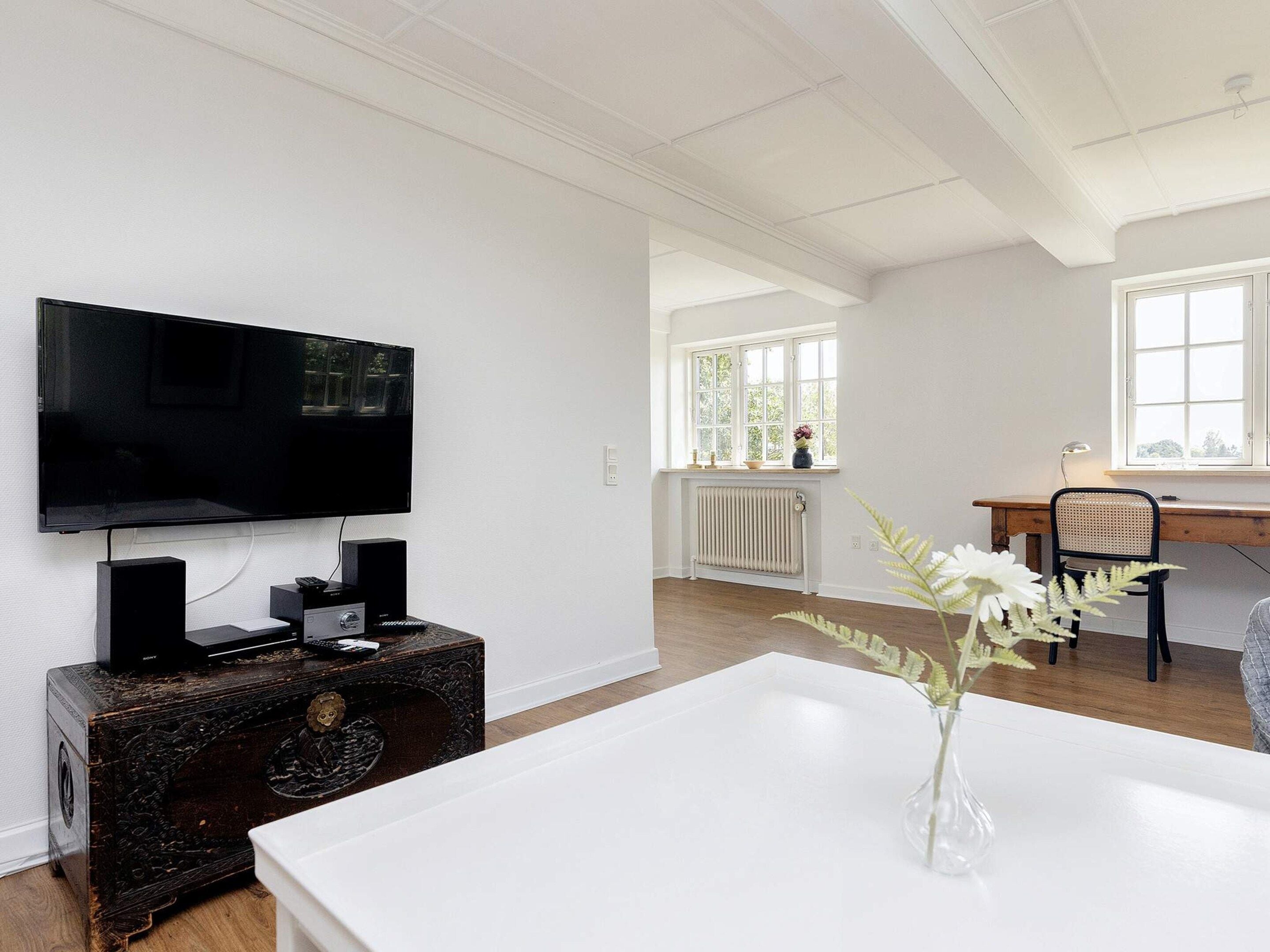 A modern living room with a flat-screen TV, a wooden chest of drawers, a white table, and a window with a view of greenery.