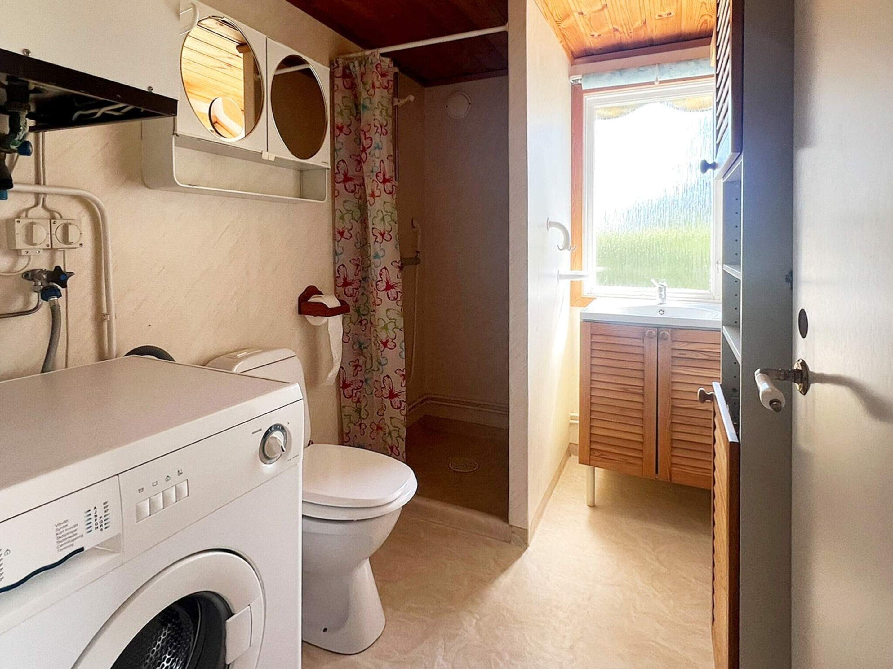 A compact bathroom with a washing machine, toilet, and a window with a view of greenery.