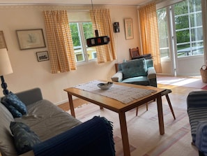 A living room with a sofa, armchair, table, and a view through a window with curtains.