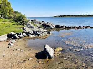 A rocky shoreline with clear water, a grassy area, and a distant hill. - 6 Person Holiday Home in Ramdala (Ramdala)