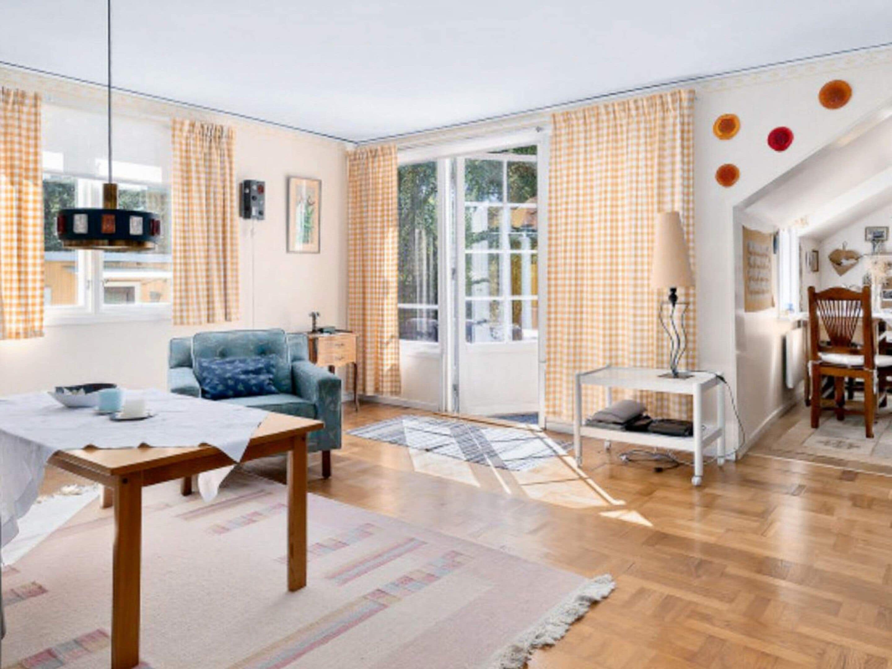 Ein geräumiges Wohnzimmer mit einer Couch, einem Esstisch und einem Sessel. Es gibt ein Fenster mit Vorhängen, eine Lampe und einen Blick ins Freie.