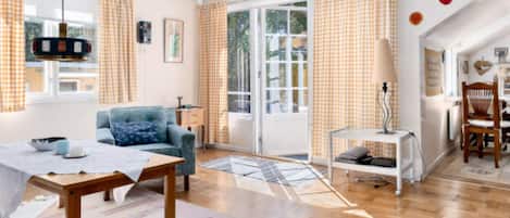 A spacious living room with a sofa, a dining table, and a chair. There is a window with curtains, a lamp, and a view of the outside.