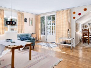 A spacious living room with a sofa, a dining table, and a chair. There is a window with curtains, a lamp, and a view of the outside.
