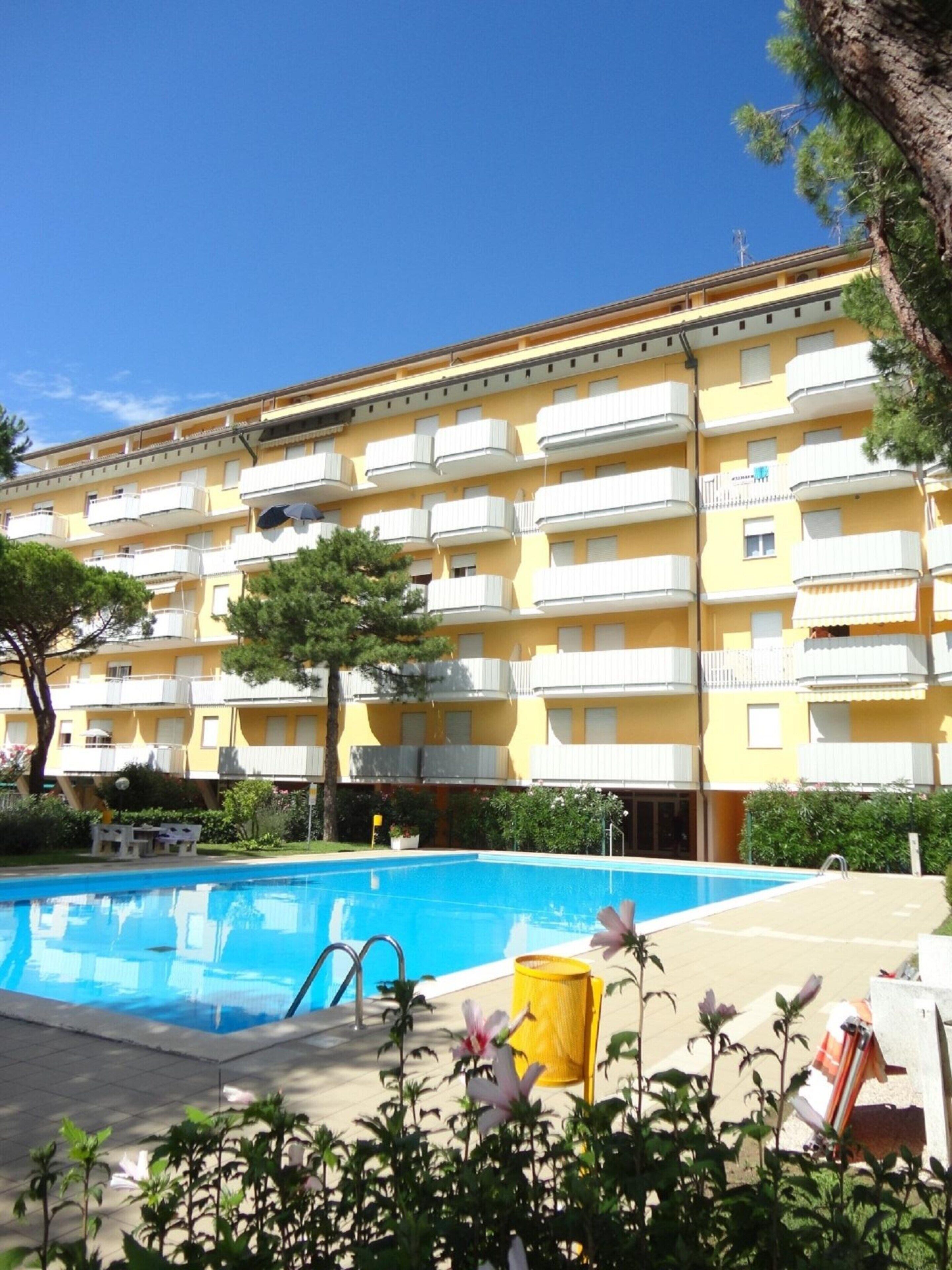 A multi-story yellow building with balconies, a swimming pool, and surrounding greenery.