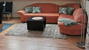 Un salon chaleureux avec un canapé incurvé, un petit pouf noir et un grand lampadaire. Des fenêtres de toit sont situées au-dessus du canapé, et on aperçoit une chaise au fond de la pièce.