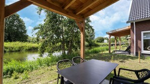 A covered outdoor area with a table and chairs, a brick building, and a body of water in the background. - Holiday Home by Lake Beach Simonshaven (Simonshaven)