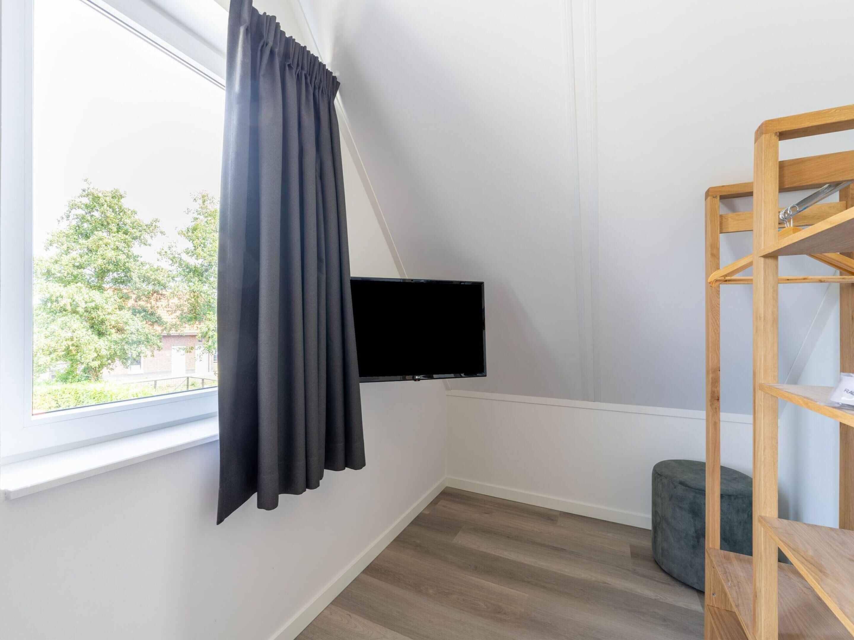 A room with a window, a black TV mounted on the wall, a wooden shelf, and a grey ottoman.