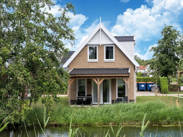 A two-story house with a sloped roof, a covered porch, and a pond in the foreground. - Holiday Home by Lake Beach Simonshaven (Simonshaven)