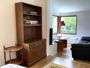 A room with a wooden cabinet, a television, a table with a lamp, a black leather sofa, and a view of greenery through a window. - 2 Person Holiday Home in Norrkoping (Norrköping)