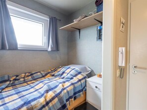 A small, neatly arranged room with a bed, a nightstand, a shelf with towels, a window with curtains, and a door with a handle. - Studio Apartment in Seaside With Panoramic Views (Middelkerke)