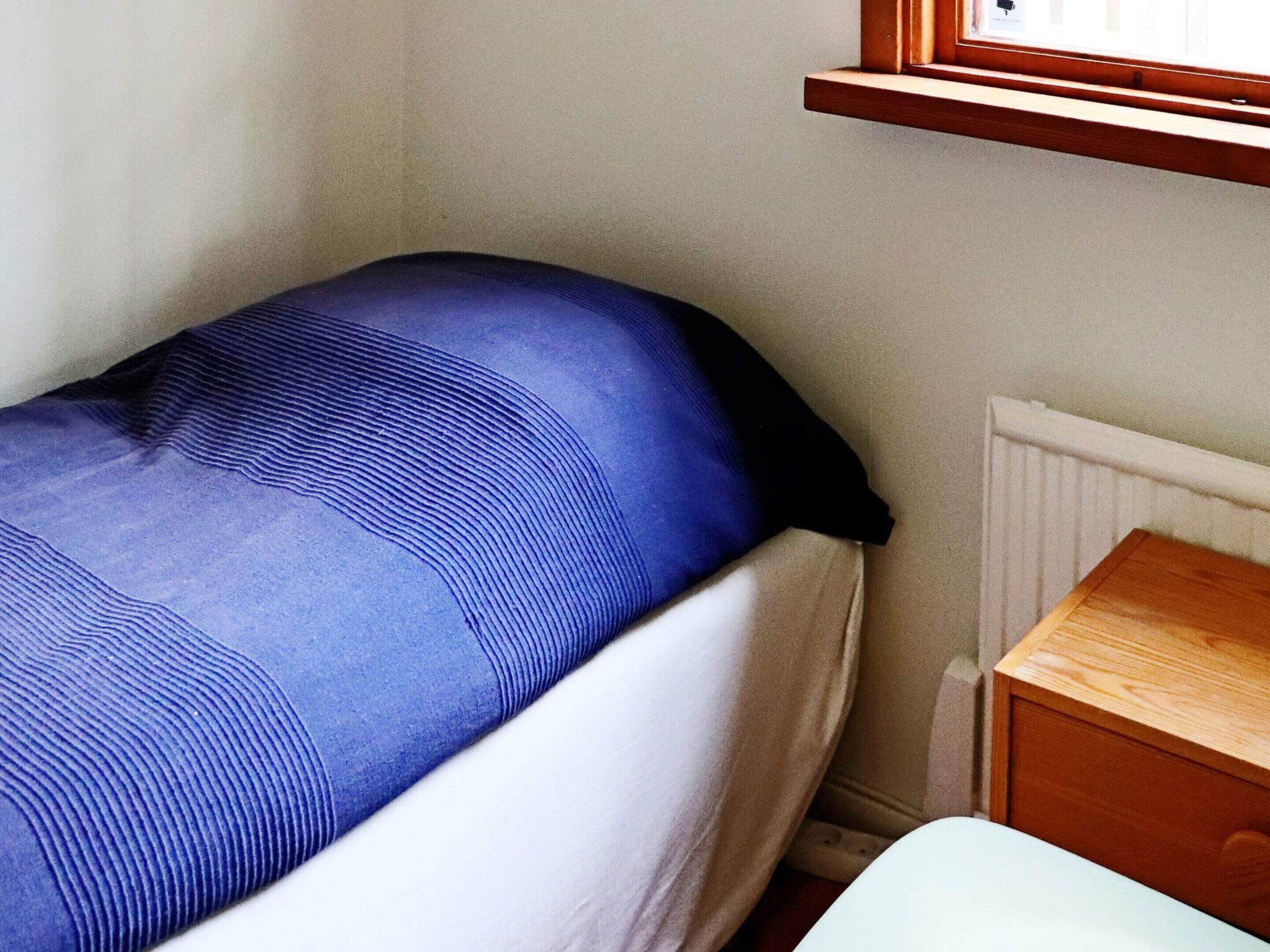Una cama individual con colchón azul y ropa de cama blanca, una mesita de noche de madera y un radiador blanco.