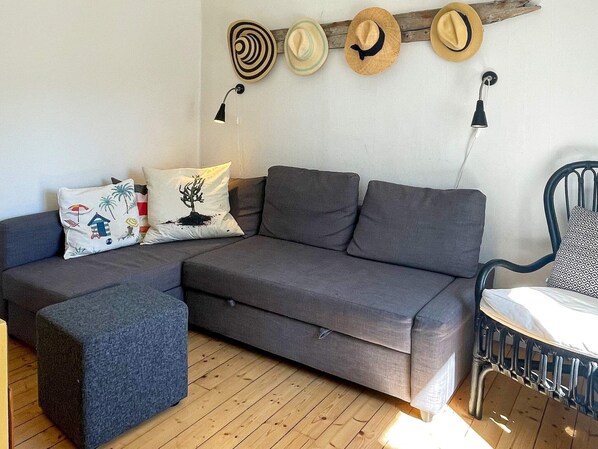 A modern living room with a grey sofa, a blue ottoman, and a black metal chair. There are three hats hanging on the wall and two wall-mounted lamps.