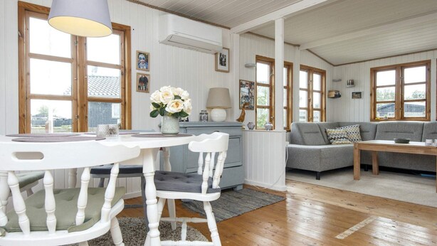 A cozy living room with a grey sofa, a white dining table with two chairs, and a kitchen area with a stove and a hanging lamp.