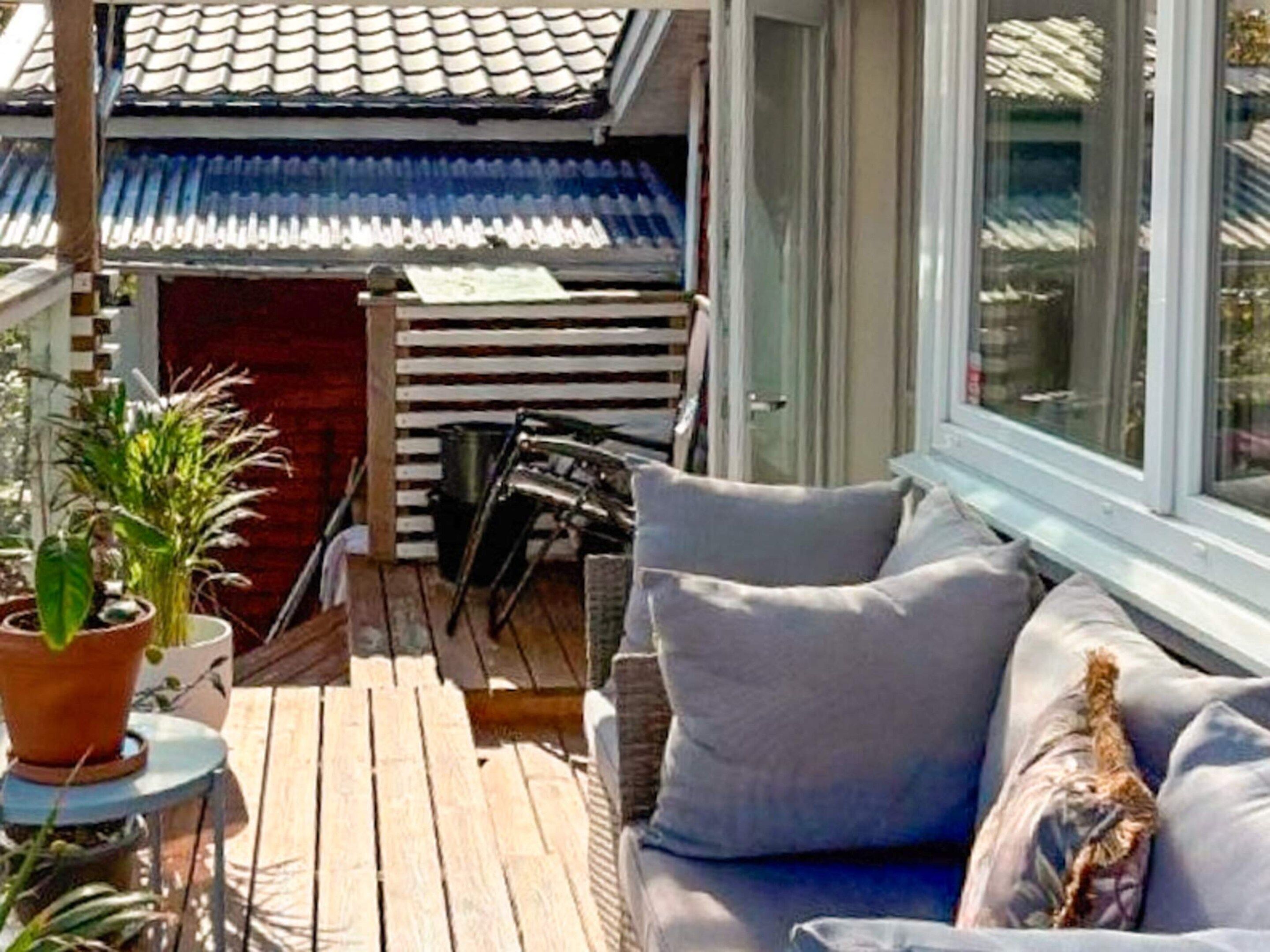 Um deck de madeira com sofá e cadeiras, plantas em vasos e vista para um edifício com telhado de azulejos.