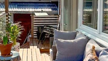 Um deck de madeira com sofá e cadeiras, plantas em vasos e vista para um edifício com telhado de azulejos.