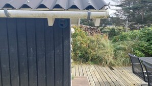 Une terrasse en bois avec une rambarde en métal, un espace couvert doté d’un toit en tôle ondulée et un mur de pierre en arrière-plan.