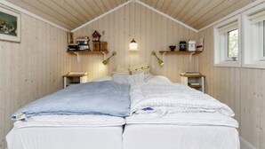 Une pièce avec un plafond en pente en bois, un lit avec une literie blanche, deux tables de chevet, une étagère murale avec des livres et des objets décoratifs, et une fenêtre laissant entrer la lumière du jour.