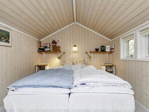 Une pièce avec un plafond en pente en bois, un lit avec une literie blanche, deux tables de chevet, une étagère murale avec des livres et des objets décoratifs, et une fenêtre laissant entrer la lumière du jour.