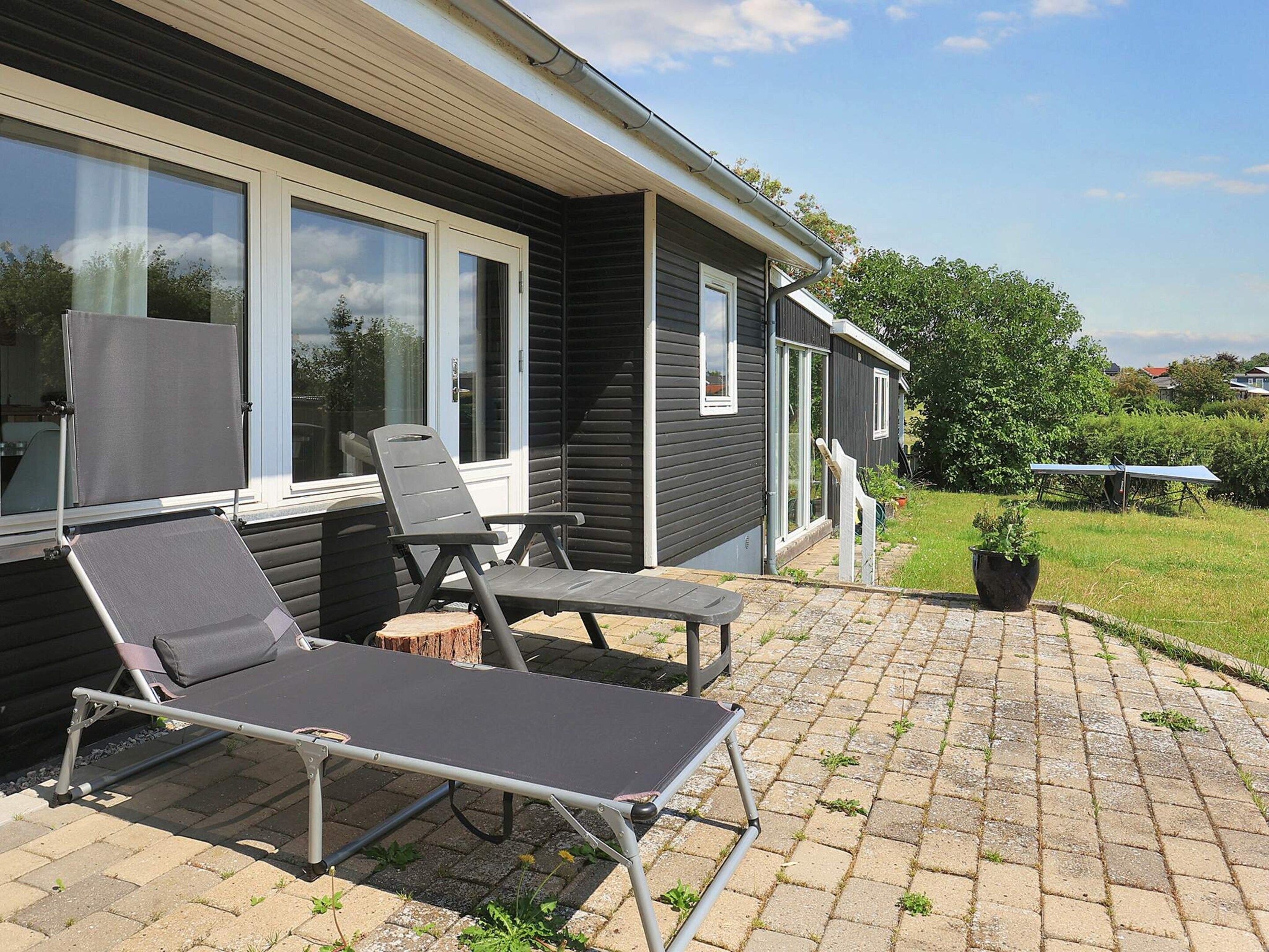 A patio with two reclining chairs and a table, a potted plant, and a house with large windows and a covered porch.