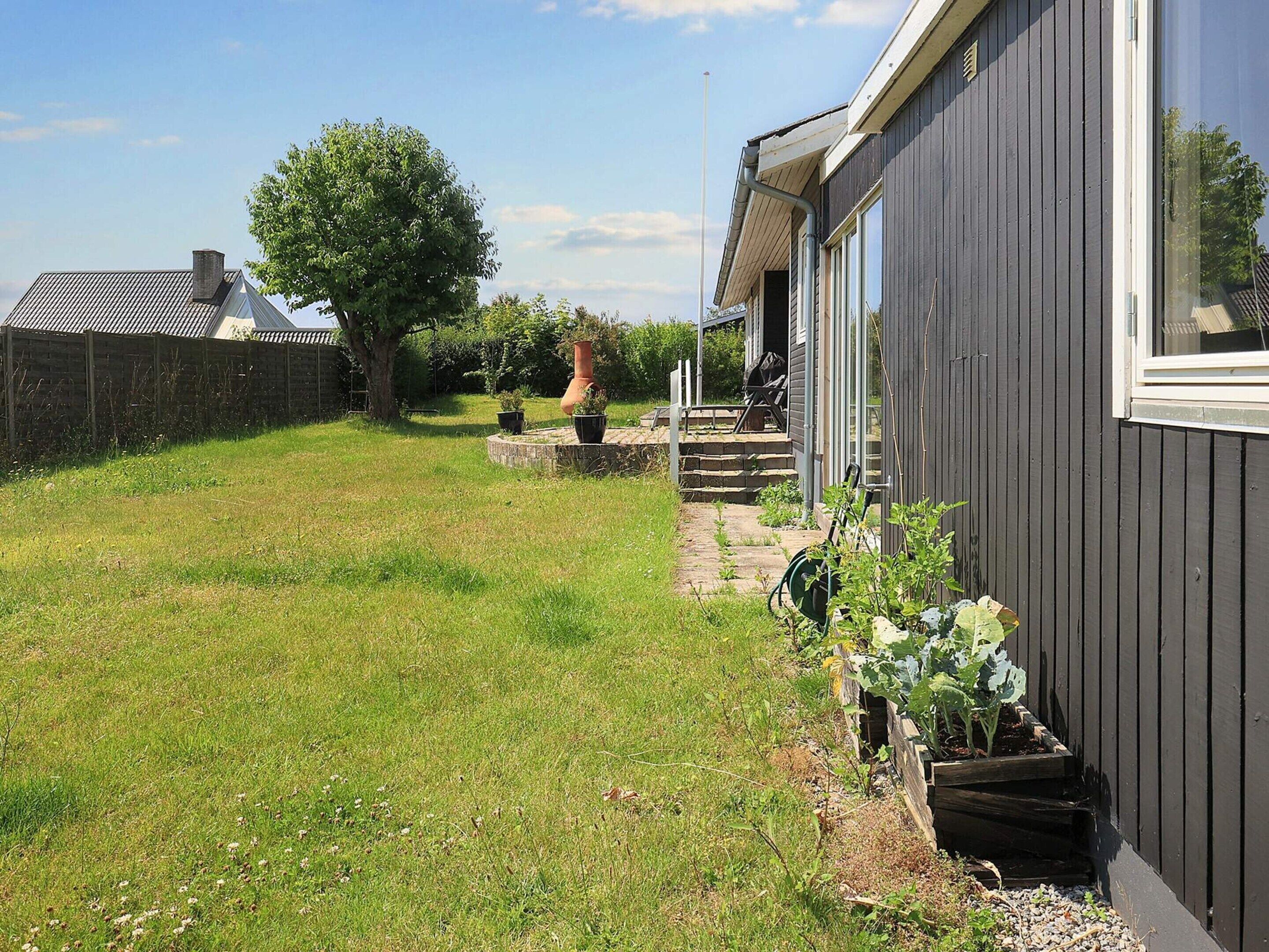 Il s’agit d’une propriété résidentielle avec un chemin pavé menant à une terrasse en bois, un grand arbre et un espace jardin agrémenté de plantes en pot.