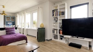 Ein modernes Wohnzimmer mit Flachbildfernseher, Bücherregal, Essbereich und großen Fenstern.