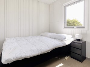 A single bed with a white floral duvet and pillows, a bedside table with a lamp, and a window with a view of greenery. - 4 Person Holiday Home in Karrebaeksminde (Karrebæksminde)