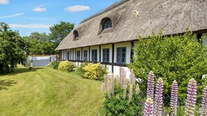 A traditional thatched-roof house with a white exterior and dark trim, surrounded by lush greenery and flowering plants. - 8 Person Holiday Home in Hadsund (Hadsund)