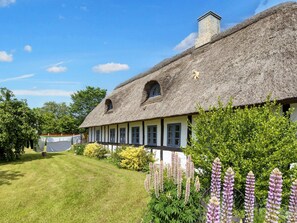 A traditional thatched-roof house with a white exterior and dark trim, surrounded by lush greenery and flowering plants. - 8 Person Holiday Home in Hadsund (Hadsund)