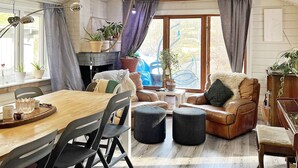 A cozy living room with a wooden dining table, leather sofa, and ottomans. There are potted plants, a fireplace, and large windows with curtains. - 8 Person Holiday Home in Ingaro (Ingaro)