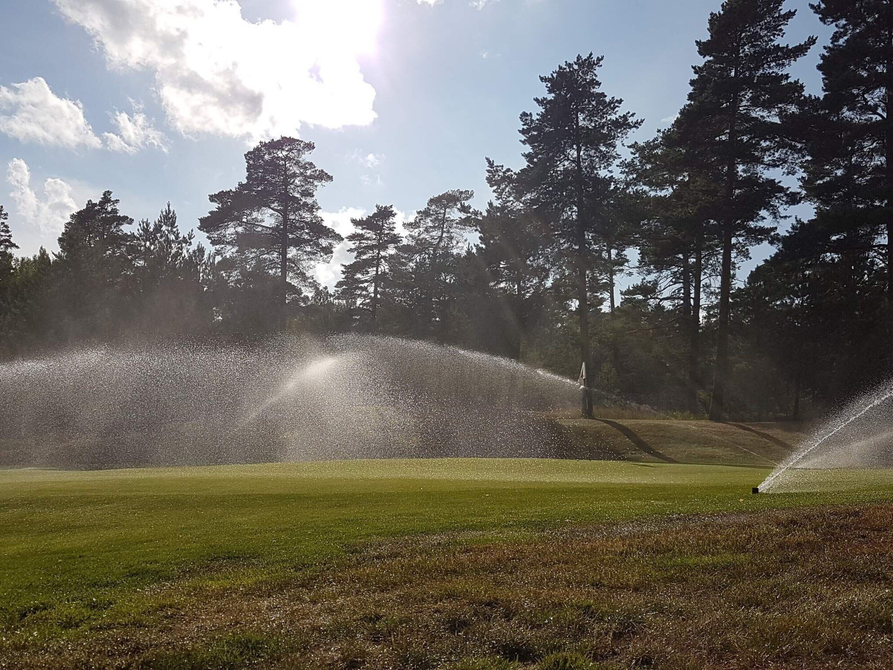 A golf course with sprinklers watering the grass, surrounded by trees and a clear sky.