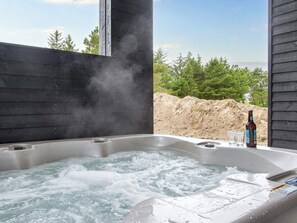 Un bain à remous avec jets de vapeur, une bouteille et des verres sur une terrasse en bois offrant une vue sur les arbres et une zone sablonneuse.