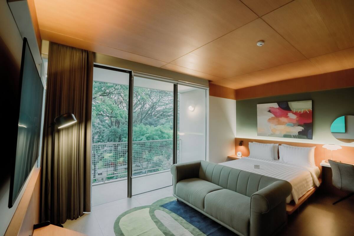 Quarto de hotel moderno com uma cama grande, um sofá, uma escrivaninha e vista para o verde através de portas deslizantes.