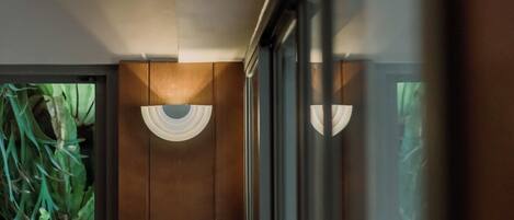 A hallway with wooden paneling, a window with a view of green foliage, and wall-mounted lighting fixtures.