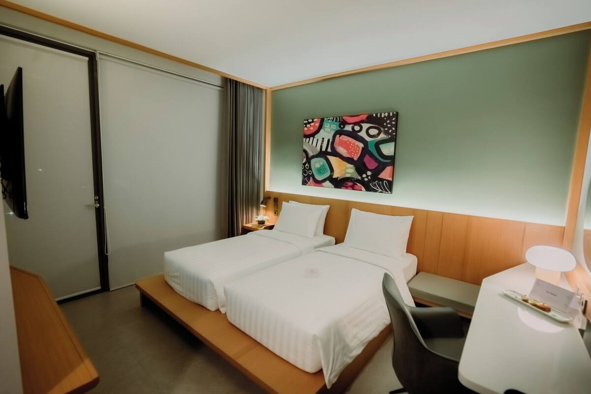 Quarto de hotel com duas camas, uma escrivaninha, uma cadeira, uma televisão e um quadro na parede.