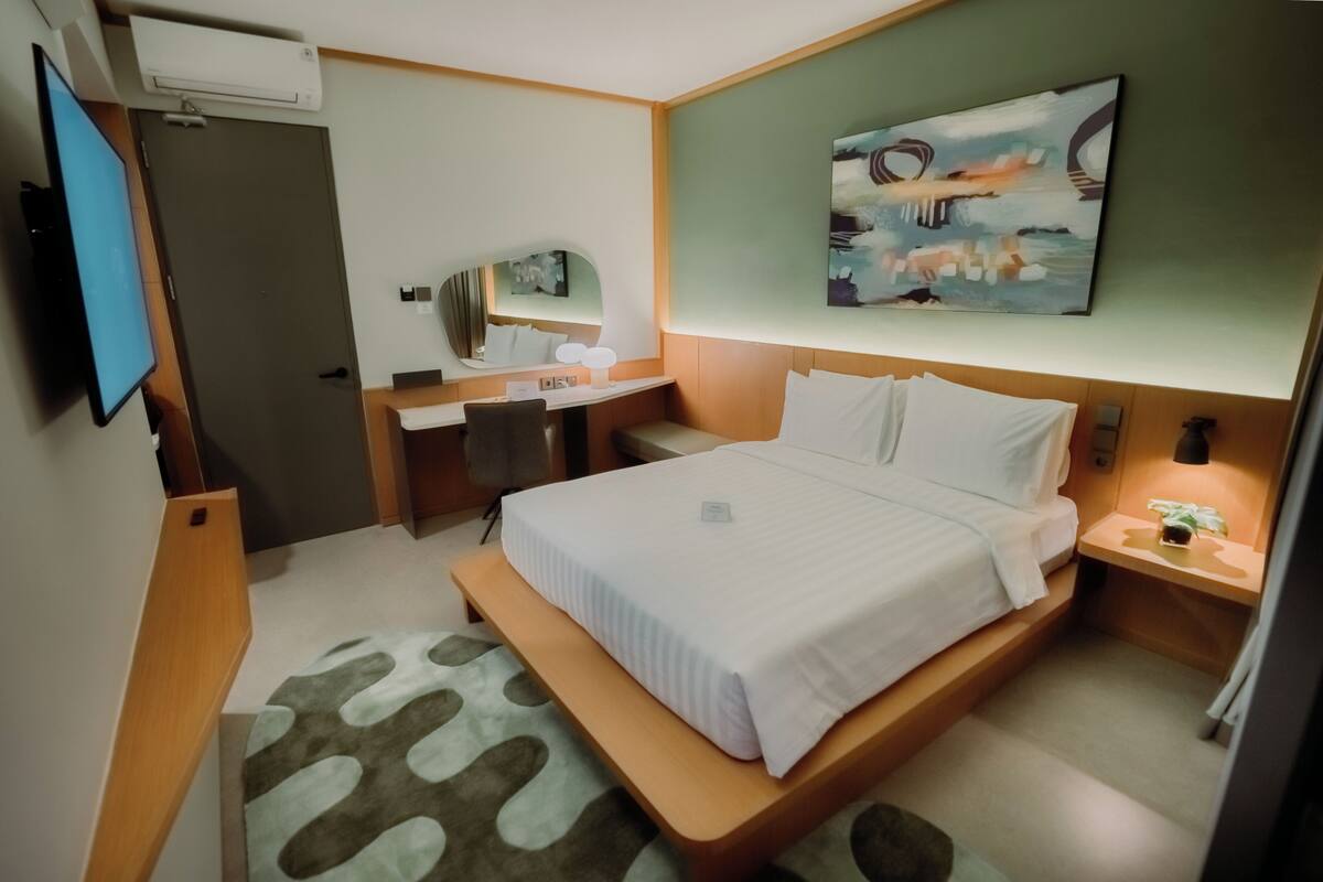 Quarto de hotel com cama, escrivaninha, espelho e televisão presa à parede.