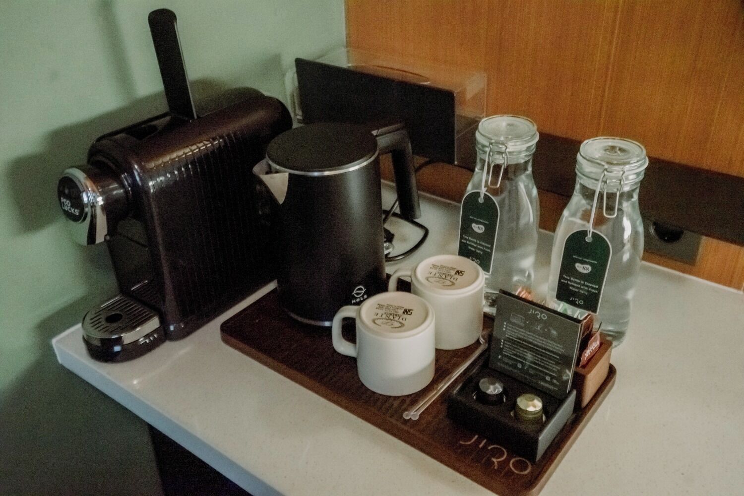 Een kitchenette met een koffiezetapparaat, waterkoker, twee mokken en een fles water op een dienblad.