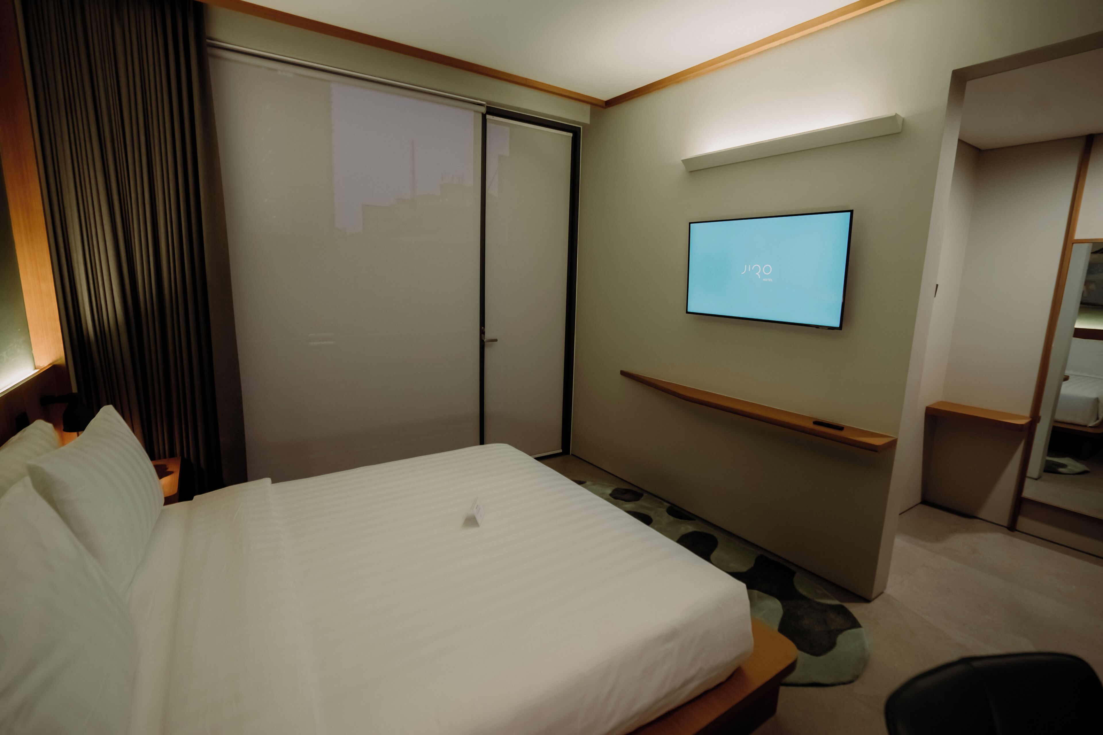 A hotel room with a bed, a TV mounted on the wall, a wardrobe, and a desk.