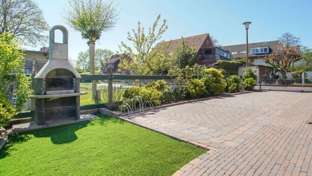 A paved patio area with a stone barbecue grill, surrounded by a green lawn and a metal fence. - At Home Comfortable Holiday Residence (Borkum)